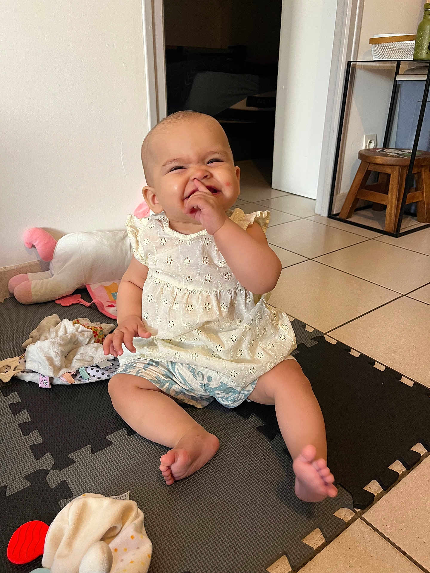 Eliana a rejoint le concours — aidez-le/la à gagner de superbes lots ! baby, child, sitting, play_mat, indoor, white_dress, shorts, toys, floor, happy, smiling, finger_in_nose, cute, infant, footwear_free, home, furniture, wooden_stool, door, tile_floor