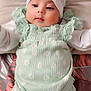 adorable, baby, bow, child, cute, face, floral_pattern, green_clothing, headband, indoors, infant, long_sleeves, lying_down, person, portrait, small, smiling, soft_surface, white_clothing, young_child