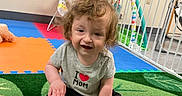 Enzo is registered to the contest to win money with this photo: toddler, child, curly_hair, grey_shirt, black_shorts, sitting, green_carpet, play_mat, playpen, toys, indoor, smiling, baby, colorful, wall_decor, photographs, flooring, happy, person, young_child