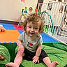toddler, child, curly_hair, grey_shirt, black_shorts, sitting, green_carpet, play_mat, playpen, toys, indoor, smiling, baby, colorful, wall_decor, photographs, flooring, happy, person, young_child