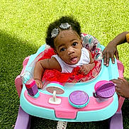 Niamani is registered to the contest to win money with this photo: adult_hand, baby, child, closeup, colorful, curious, cute, expression, grass, hair_accessories, hands, outdoor, playtime, red_tutu, smiling, summer, sunlight, toddler, toy, walker