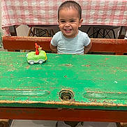 Enzo participe au concours pour gagner de l'argent avec cette photo : toddler, child, smiling, toy_car, table, wooden_table, green_table, bench, indoor, furniture, checkered_tablecloth, happy, person, playful, cute, face, short_hair, home, chair, plastic_bottle
