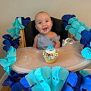 Easton joined the competition — help win amazing prizes! baby, high_chair, birthday, cupcake, smiling, blue_garland, decoration, celebration, infant, happy, face, person, wooden_floor, table, clothing, indoor, party, colorful, food, seat