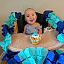 baby, high_chair, birthday, cupcake, smiling, blue_garland, decoration, celebration, infant, happy, face, person, wooden_floor, table, clothing, indoor, party, colorful, food, seat