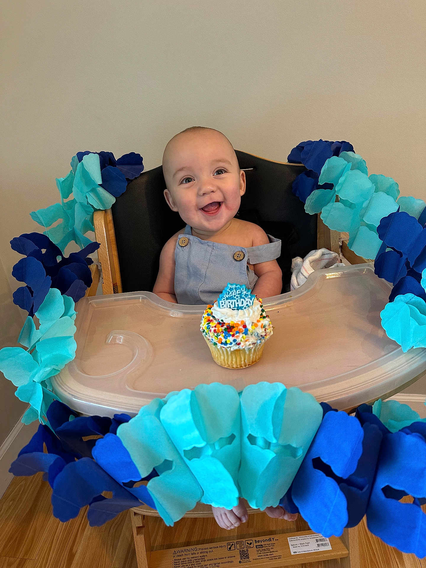 Easton joined the competition — help win amazing prizes! baby, high_chair, birthday, cupcake, smiling, blue_garland, decoration, celebration, infant, happy, face, person, wooden_floor, table, clothing, indoor, party, colorful, food, seat