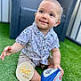 baby, background, child, face, foot, grass, green, hand, happy, month_marker, outdoor, patterned_clothing, rugby_ball, shirt, shorts, sitting, smile, toy, wicker_chair, wooden_sign