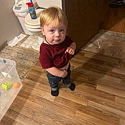 Huxson is registered to the contest to win money with this photo: child, toddler, bathroom, toilet, wooden_floor, sippy_cup, blond_hair, necklace, jeans, socks, standing, looking_at_camera, toy_ball, plastic_container, tile_floor, candid, person, trash_can, indoor, curious_expression