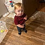child, toddler, bathroom, toilet, wooden_floor, sippy_cup, blond_hair, necklace, jeans, socks, standing, looking_at_camera, toy_ball, plastic_container, tile_floor, candid, person, trash_can, indoor, curious_expression
