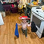 child, toddler, hat, kitchen, stove, hardwood_floor, barefoot, cloth, diaper, countertop, fruits, mango, orange, pan, chair, bag, storage_bins, blanket, messy_room, playful