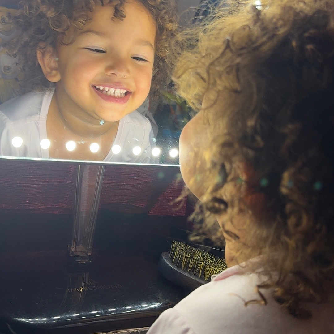 Lelianna is registered to the contest to win money with this photo: child, close_up, curly_hair, cute, expression, face, glow, hair_brush, happy, indoor, joy, lights, mirror, portrait, reflection, smiling, table, vanity_mirror, white_shirt, young_child