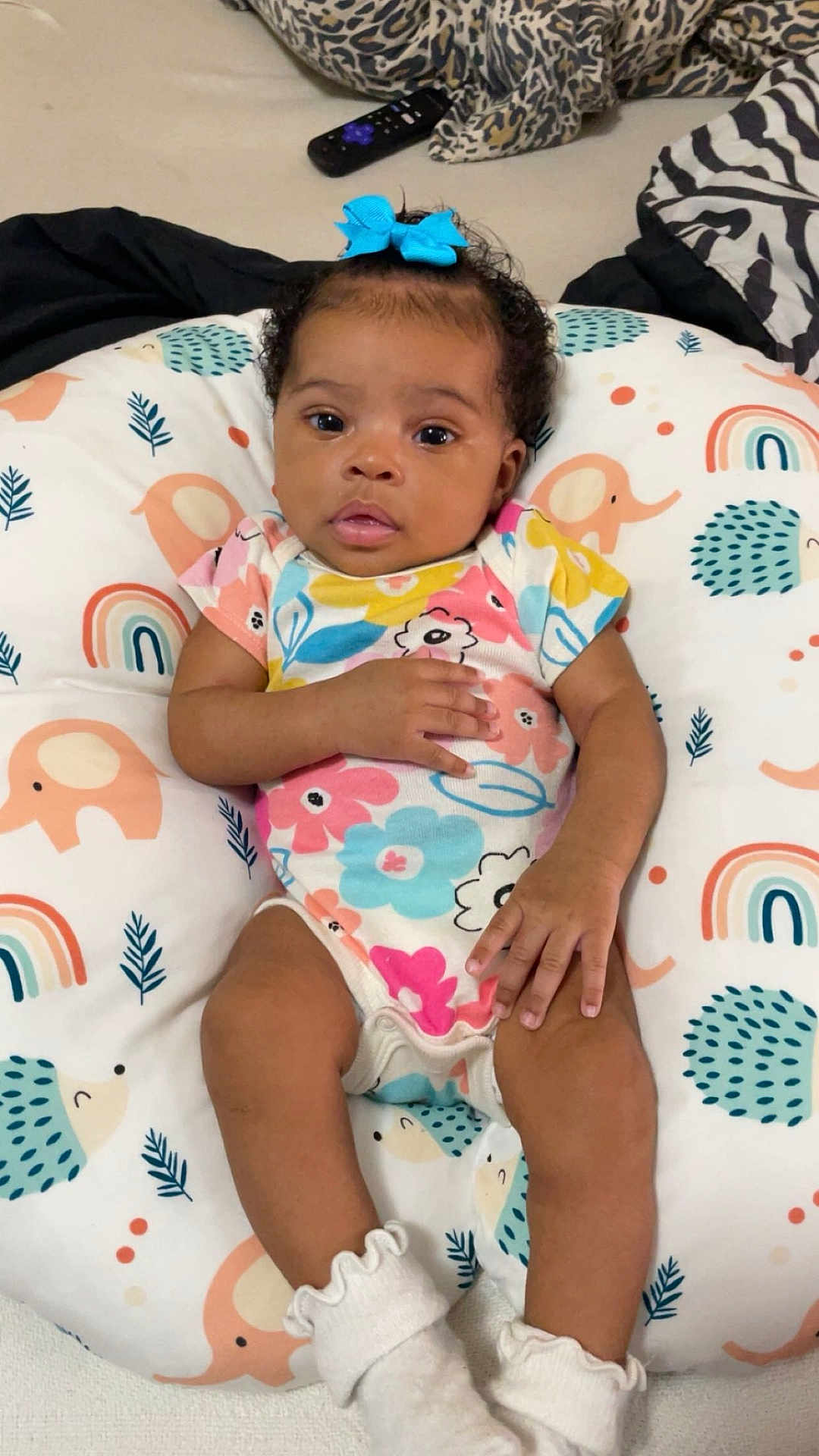 Neorah is registered to the contest to win money with this photo: baby, infant, child, bow, blue_bow, onesie, floral_pattern, socks, cushion, animal_pattern, rainbow_pattern, face, hands, legs, curious, indoor, soft_toys, colorful, lying_down, portrait