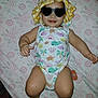 baby, infant, sunglasses, hat, onesie, patterned_clothing, smile, lying_down, bed_sheet, floral_pattern, cute, child, person, cheerful, indoor, skin, legs, arms, headwear, accessory