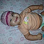 baby, infant, smiling, hat, knitted_hat, tshirt, diaper, lying_down, bed_sheet, floral_pattern, toy, drool, cute, child, happy, fists, pink, peach, indoor, person