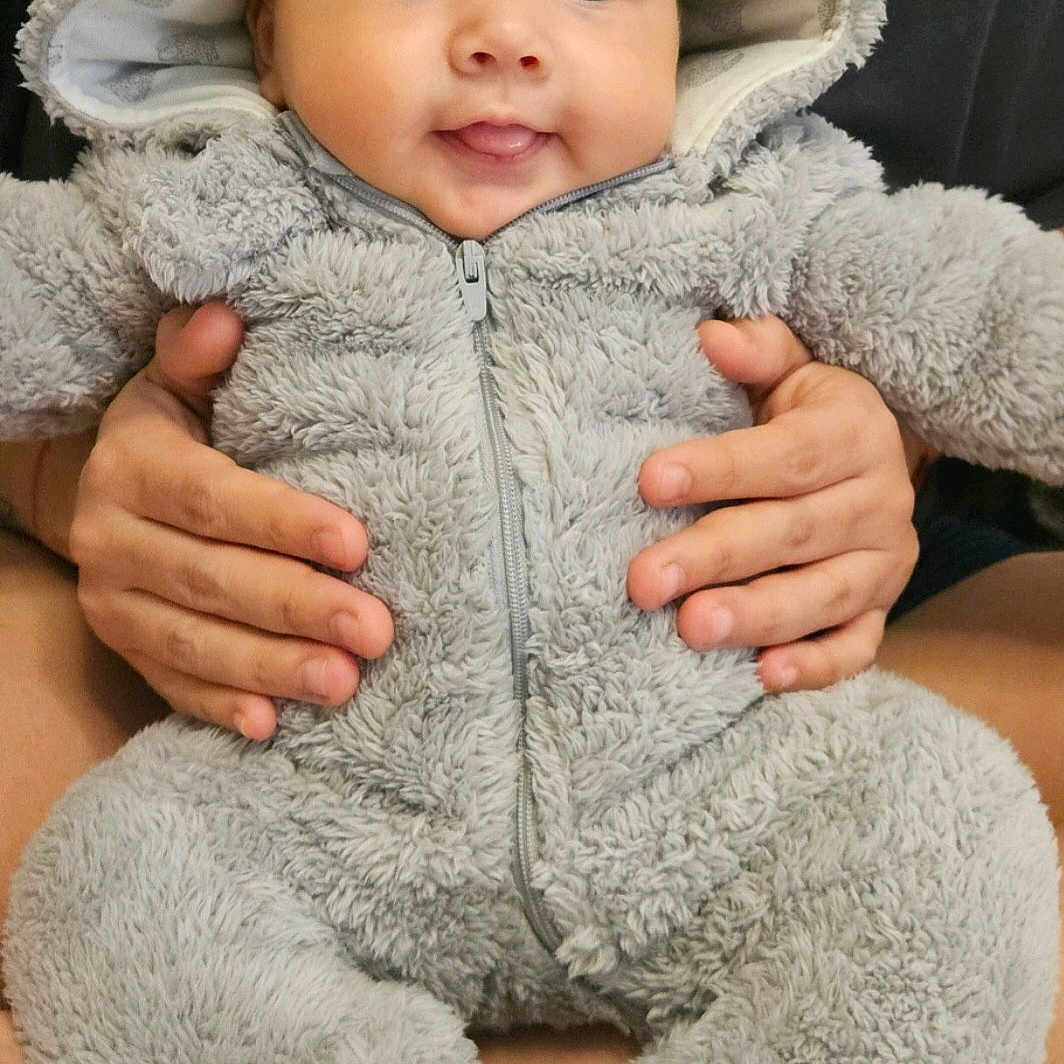 Hazel is registered to the contest to win money with this photo: baby, child, clothing, cozy, cute, cute_outfit, ears, face, fluffy, gray, hands, happy, holding, hood, indoor, infant, onesie, portrait, smiling, toddler