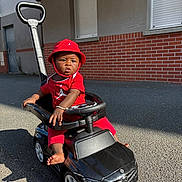 Ismaël participe au concours pour gagner de l'argent avec cette photo : toddler, child, red_clothing, red_hat, toy_car, black_toy_car, outdoor, asphalt, sunlight, shadow, brick_wall, window, building, steering_wheel, barefoot, bracelet, serious_expression, daytime, person, playtime