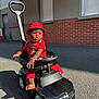 toddler, child, red_clothing, red_hat, toy_car, black_toy_car, outdoor, asphalt, sunlight, shadow, brick_wall, window, building, steering_wheel, barefoot, bracelet, serious_expression, daytime, person, playtime