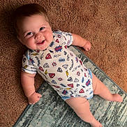 Dallas is registered to the contest to win money with this photo: baby, child, smiling, onesie, superhero, carpet, rug, indoor, cute, happy, infant, person, face, skin, hand, foot, clothing, laying, cheerful, portrait