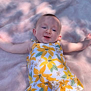 Nevaeh is registered to the contest to win money with this photo: baby, child, infant, smiling, face, arms, dress, floral_pattern, blanket, soft_texture, outdoor, sunlight, shadow, happy, cute, lying_down, green_pants, portrait, young_child, person