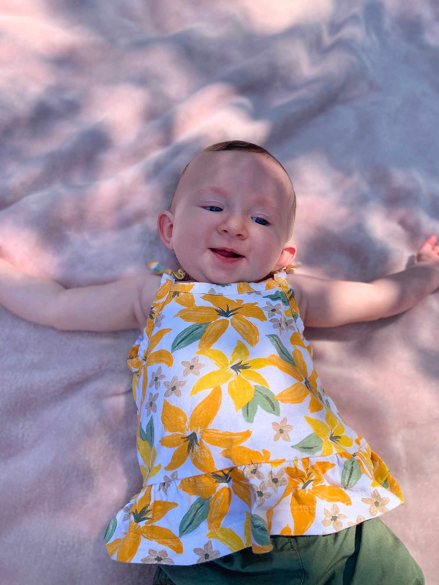Nevaeh is registered to the contest to win money with this photo: baby, child, infant, smiling, face, arms, dress, floral_pattern, blanket, soft_texture, outdoor, sunlight, shadow, happy, cute, lying_down, green_pants, portrait, young_child, person