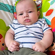 William is registered to the contest to win money with this photo: animal_pattern, baby, child, chubby_cheeks, closeup, colorful_baby_seat, comfortable, cute, fists, green_pants, indoor, infant, leaf_pattern, playful, portrait, serious_expression, sitting, soft_lighting, striped_shirt, young_child