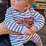 baby, child, striped_clothing, sitting, adult_hand, outdoor, stroller, wicker_basket, curious, expression, sunlight, portrait, infant, casual, jeans, hands, flooring, toy, person, daylight