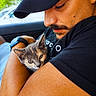 man, kitten, cat, pet, animal, human, face, arm, watch, black_tshirt, cap, car_interior, window, closeup, affection, caring, cute, young, indoor, portrait