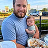 man, baby, child, beard, smile, food, meal, drink, outdoor, table, plate, car, greenery, sky, person, clothing, face, holding, restaurant, daytime