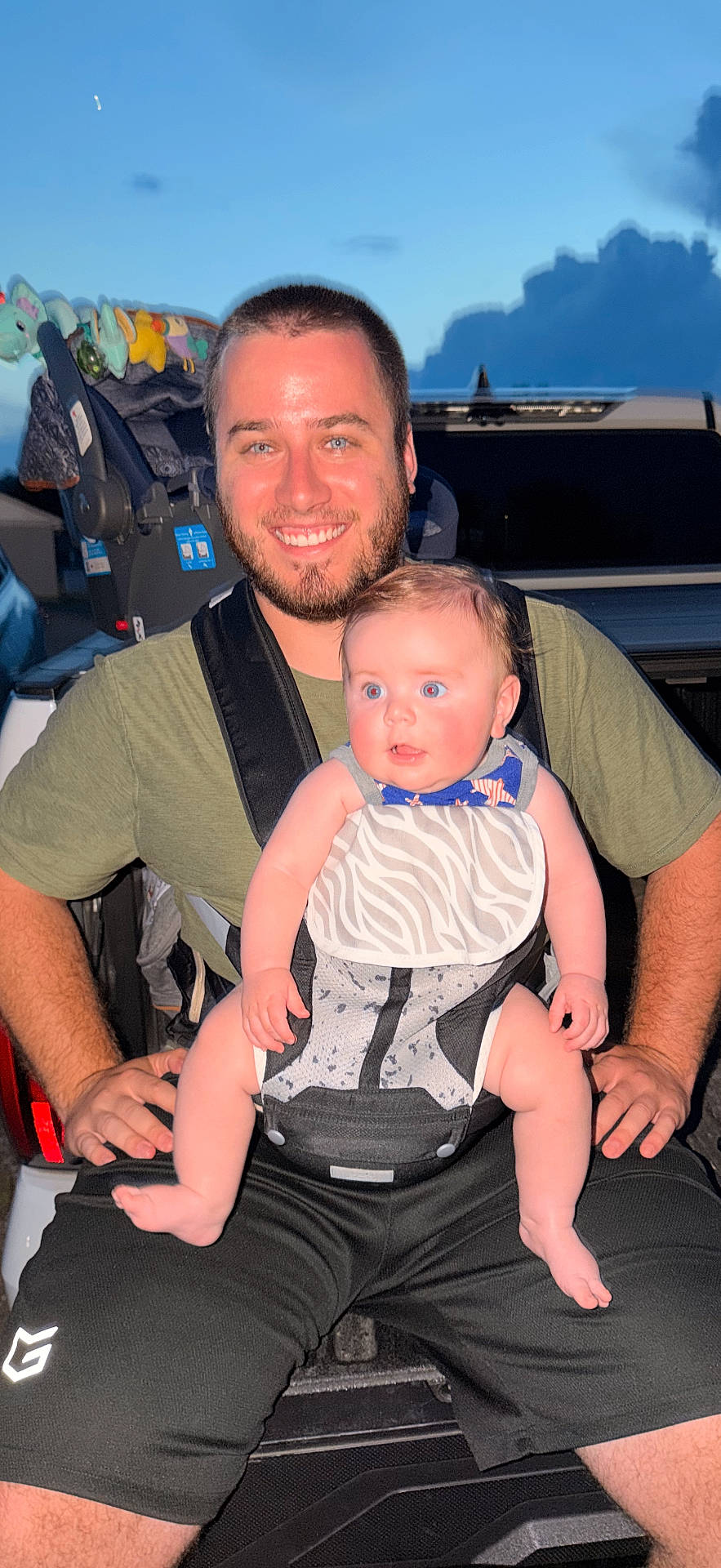Kameron is registered to the contest to win money with this photo: baby, beard, bib, carrier, casual, child, clothing, clouds, daytime, family, happy, man, outdoor, person, portrait, shorts, sitting, sky, smiling, vehicle