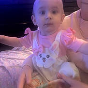 Luna is registered to the contest to win money with this photo: baby, child, person, pink_clothing, bunny_design, adult_hand, glasses, indoor, fabric, holding, face, portrait, cute, infant, sitting, soft_lighting, blurred, purple_hue, short_hair, young_child