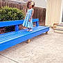 child, girl, blue_dress, flower, curly_hair, bench, outdoor, backyard, fence, house, concrete, smile, standing, plant, shoes, daylight, sidewalk, portrait, happy, casual
