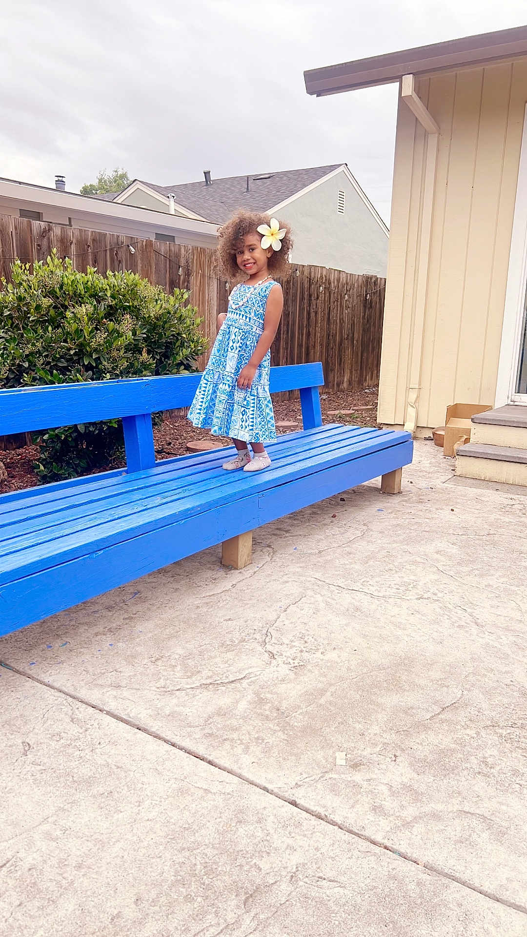 Adi Sera is registered to the contest to win money with this photo: child, girl, blue_dress, flower, curly_hair, bench, outdoor, backyard, fence, house, concrete, smile, standing, plant, shoes, daylight, sidewalk, portrait, happy, casual