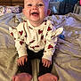 baby, smiling, heart_pattern, sweater, sitting, bed, blanket, indoor, child, happy, cute, toothless_grin, person, feet, hands, wooden_drawers, room, cozy, casual_clothing, portrait