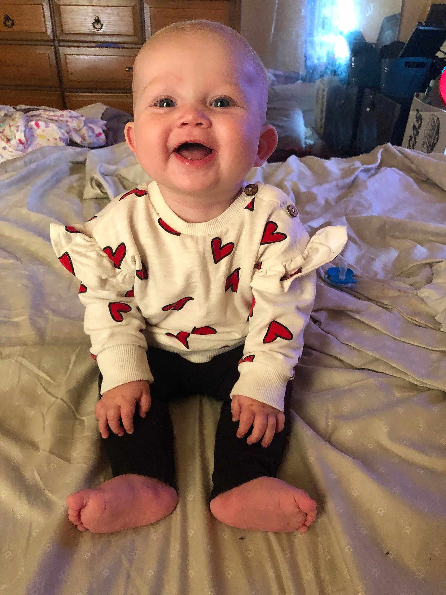 Delilah joined the competition — help win amazing prizes! baby, smiling, heart_pattern, sweater, sitting, bed, blanket, indoor, child, happy, cute, toothless_grin, person, feet, hands, wooden_drawers, room, cozy, casual_clothing, portrait