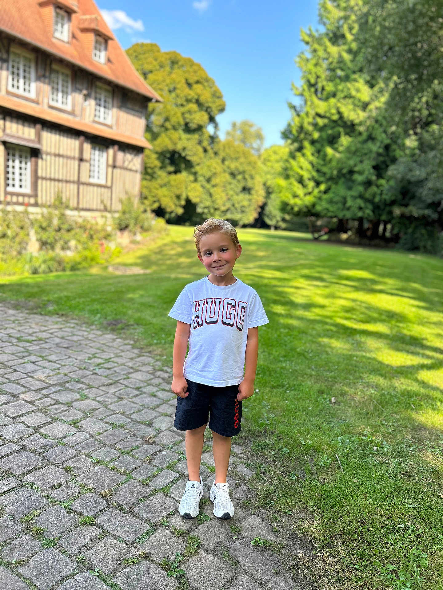 Isaac a rejoint le concours — aidez-le/la à gagner de superbes lots ! child, boy, smiling, outdoor, grass, tree, building, cobblestone, path, sky, sunny, daylight, casual_clothing, white_shirt, shorts, sneakers, nature, portrait, standing, greenery