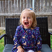 Braeleigh is registered to the contest to win money with this photo: child, toddler, smiling, laughing, dress, patterned_clothing, deer_pattern, blue_dress, hair_tie, outdoor, chair, woven_chair, fence, grass, nature, happy, cute, sitting, person, portrait