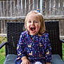 child, toddler, smiling, laughing, dress, patterned_clothing, deer_pattern, blue_dress, hair_tie, outdoor, chair, woven_chair, fence, grass, nature, happy, cute, sitting, person, portrait