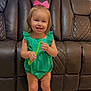 toddler, child, smiling, green_outfit, pink_bow, toy, indoor, leather_couch, flooring, sandals, happy, person, small_child, cute, playful, home, portrait, standing, face, furniture