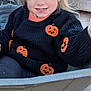 child, girl, sweater, pumpkin, halloween, wheelbarrow, outdoor, blonde_hair, pigtails, orange_bows, smiling, cute, fall, decorations, wooden_background, playful, young_child, knitwear, festive, costume