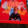 toddler, child, pumpkin, beanie, sweater, autumn, fall, outdoor, grass, illustration, red_backdrop, seasonal, cute, portrait, clothing, young_child, festive, holiday, pumpkin_pattern, casual
