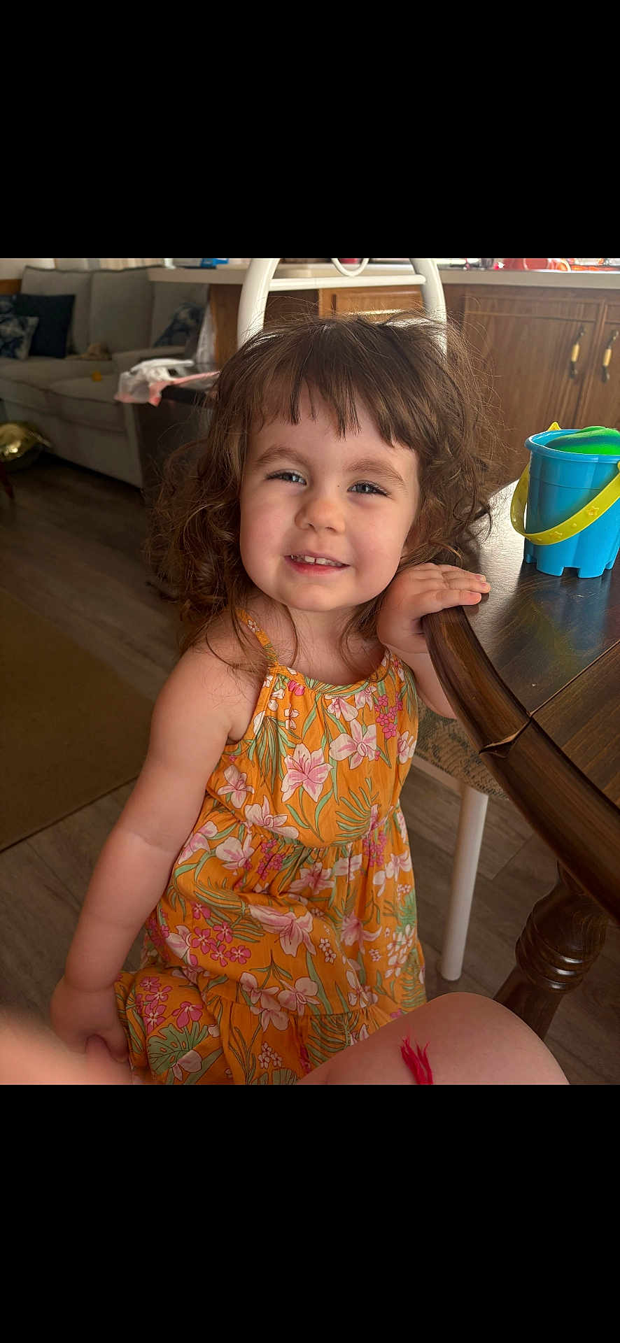 Charlie joined the competition — help win amazing prizes! child, girl, smile, curly_hair, dress, floral_pattern, table, wooden_table, toy, bucket, indoor, living_room, couch, floor, person, happy, cute, home, furniture, portrait
