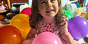 Charlie is registered to the contest to win money with this photo: child, toddler, balloons, indoor, smiling, happy, colorful, wooden_wall, dartboard, furniture, basket, toy, playful, curly_hair, sitting, dress, person, celebration, bright, cheerful