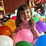 child, toddler, balloons, indoor, smiling, happy, colorful, wooden_wall, dartboard, furniture, basket, toy, playful, curly_hair, sitting, dress, person, celebration, bright, cheerful