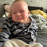 Ezra is registered to the contest to win money with this photo: baby, infant, smiling, face, clothing, striped_cardigan, pants, bed, pillow, blanket, indoor, person, cute, chubby_cheeks, portrait, happy, child, young, seated, cozy
