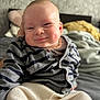baby, bed, blanket, child, chubby_cheeks, clothing, cozy, cute, face, happy, indoor, infant, pants, person, pillow, portrait, seated, smiling, striped_cardigan, young