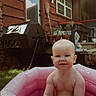 baby, infant, inflatable_pool, pink, backyard, grass, house, door, window, plants, potted_plants, compost_bin, outdoor, summer, child, smile, skin, bare_chest, daylight