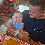 Levi joined the competition — help win amazing prizes! baby, infant, child, father, parent, smiling, happy, portrait, indoors, restaurant, fast_food, hamburger, food, drink_dispenser, red_chairs, tiled_floor, holding_baby, family, casual_photo, diner
