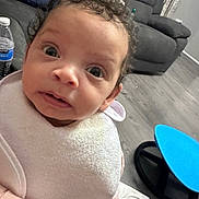 Daisy is registered to the contest to win money with this photo: baby, child, curly_hair, bib, indoor, living_room, couch, floor, toy, plastic_bottle, footwear, pink_clothing, wide_eyes, closeup, person, expression, curious, carpet, furniture, home