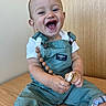 baby, child, smiling, overalls, pacifier, sitting, happy, cute, indoor, wooden_surface, wood_panel, white_shirt, blue_eyes, short_hair, infant, clothing, playful, person, portrait, joyful