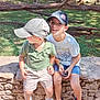 child, children, boy, cap, hat, smile, outdoor, park, stone_wall, greenery, grass, trees, sunlight, shorts, tshirt, footwear, casual, sitting, nature, playful