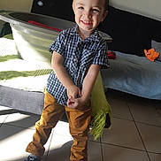 Iloan a rejoint le concours — aidez-le/la à gagner de superbes lots ! child, boy, smile, indoor, sunlight, shadow, checkered_shirt, mustard_pants, black_shoes, tile_floor, bed, blanket, laundry_basket, hair_spiky, happy, casual_clothing, home, person, floor, playful